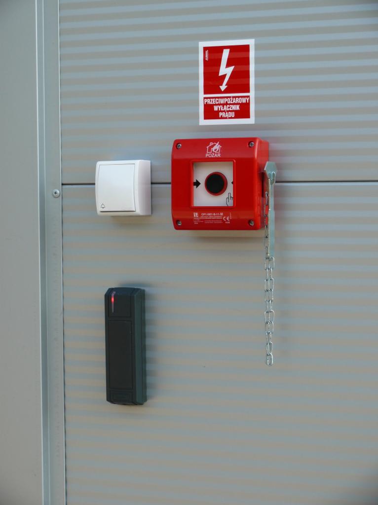 Przeciwpożarowy wyłącznik prądu w czerwonej obudowie z łańcuszkiem, biały włącznik światła i czarny czytnik kart dostępu zamontowane na szarej ścianie z poziomymi paskami.