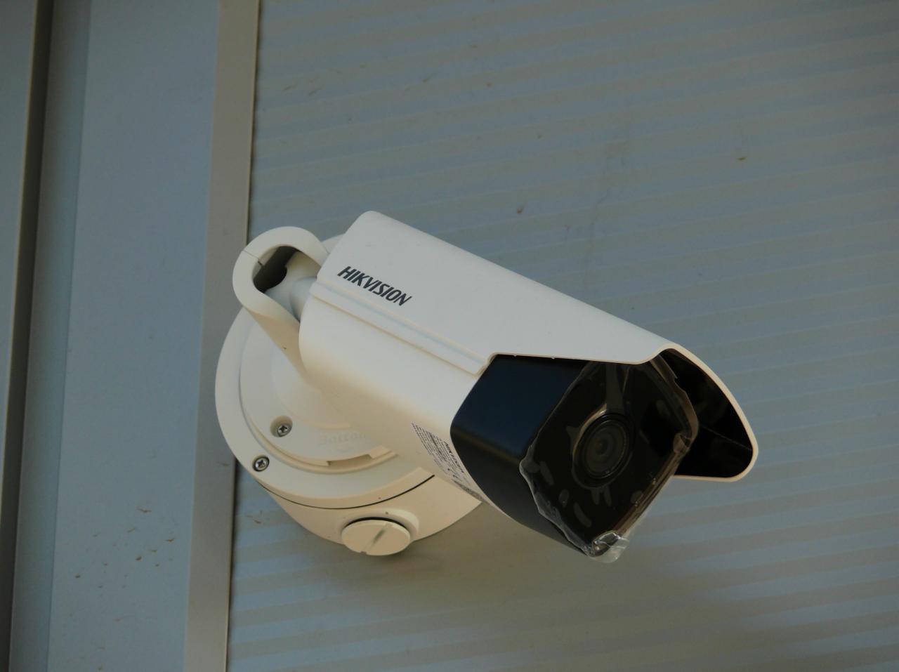 Biała kamera monitoringu Hikvision zamontowana na szarej ścianie zewnętrznej z widocznymi śrubami mocującymi i folią ochronną na obiektywie.