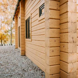 Ściana zewnętrzna domu z bali, widoczne połączenia narożne z bali, czarne ramy okienne, szara żwirowa ścieżka i jesienne drzewa w tle.