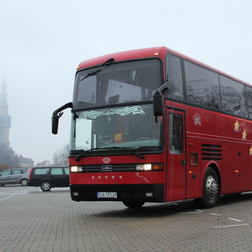 Czerwony autobus turystyczny Van Hool na parkingu z widoczną wieżą w tle, tablica rejestracyjna DOA 55528, oznaczenia Royal Class i gwiazdy.