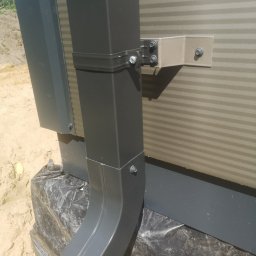 Venor - Szary rynna spustowa przymocowana do ściany z paneli warstwowych za pomocą metalowych wsporników i śrub, zabezpieczona folią budowlaną u podstawy.