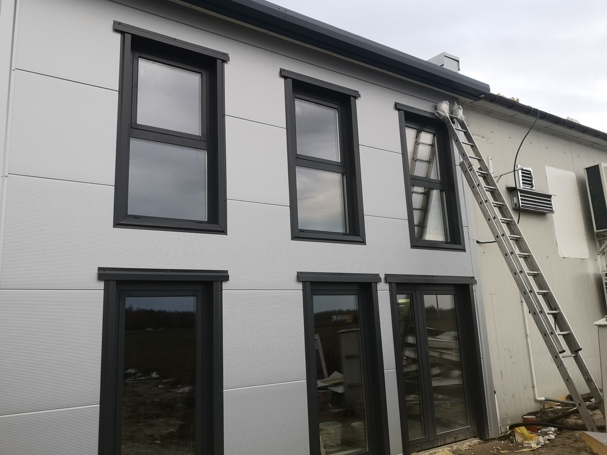 Fasada budynku obłożona jasnoszarą płytą warstwową z czarnymi ramami okiennymi i drabiną opartą o ścianę.