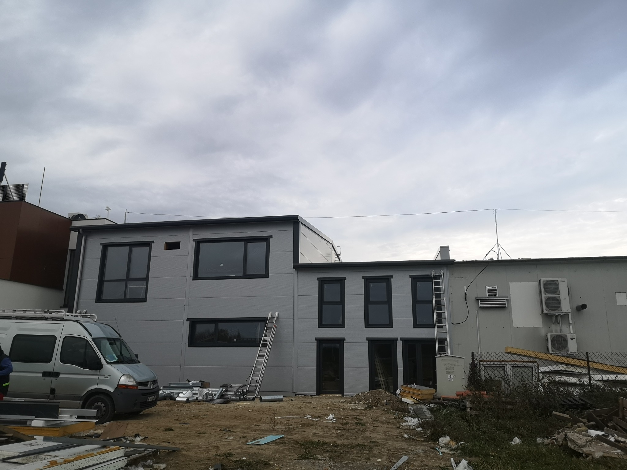 Budynek przemysłowy w trakcie montażu płyt warstwowych, widoczny srebrny van i drabina oparta o ścianę, pochmurne niebo.