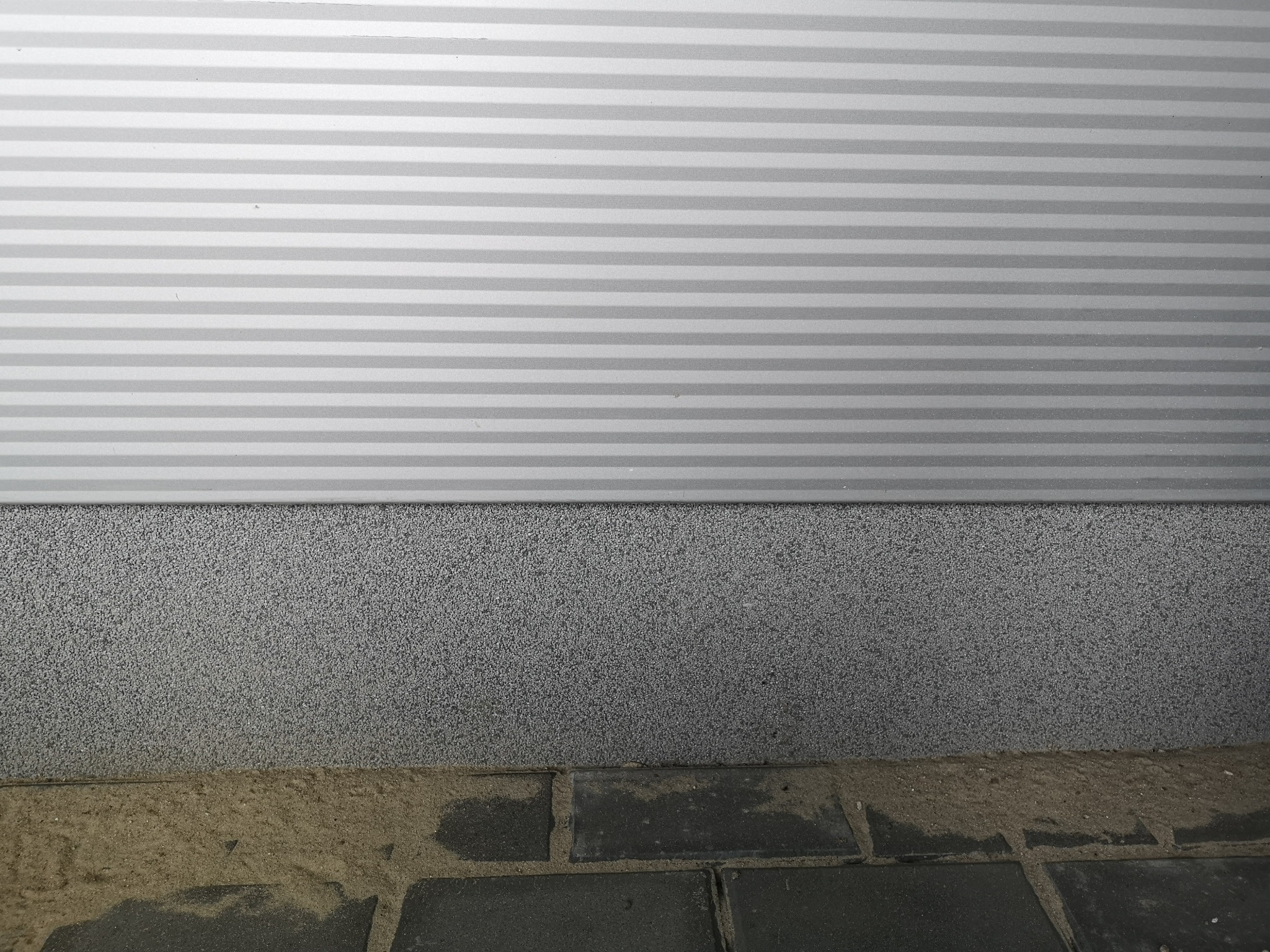 Szara, pozioma płyta warstwowa z widoczną fakturą, zakończona szarym tynkiem i fragmentem kostki brukowej z piaskiem w spoinach.
