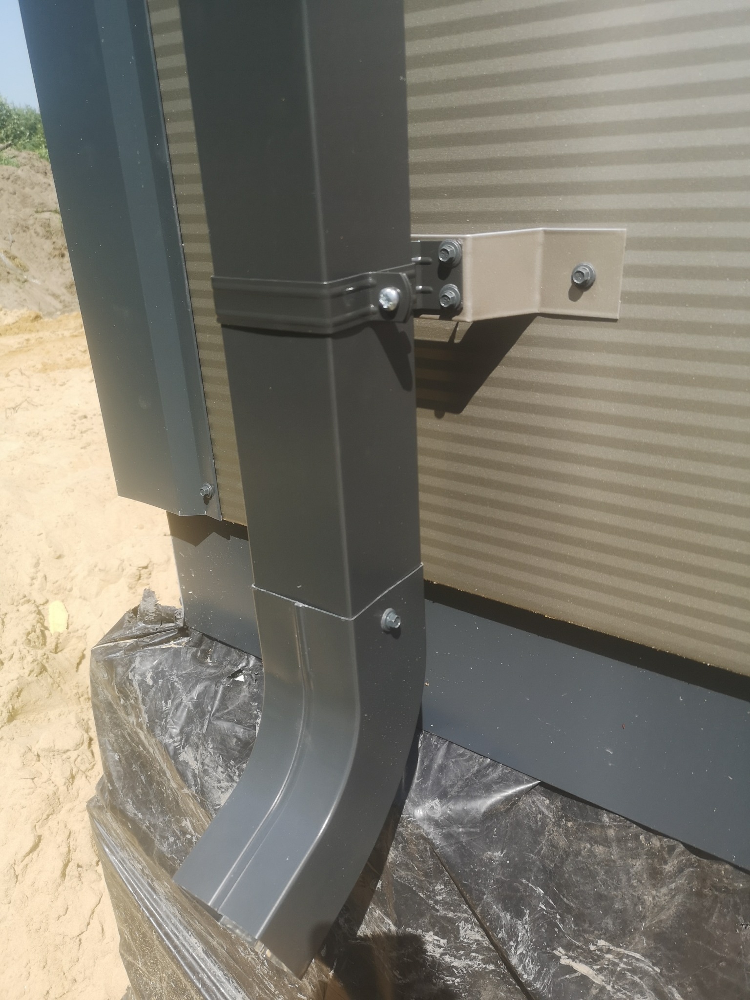 Szary rynna spustowa przymocowana do ściany z paneli warstwowych za pomocą metalowych wsporników i śrub, zabezpieczona folią budowlaną u podstawy.