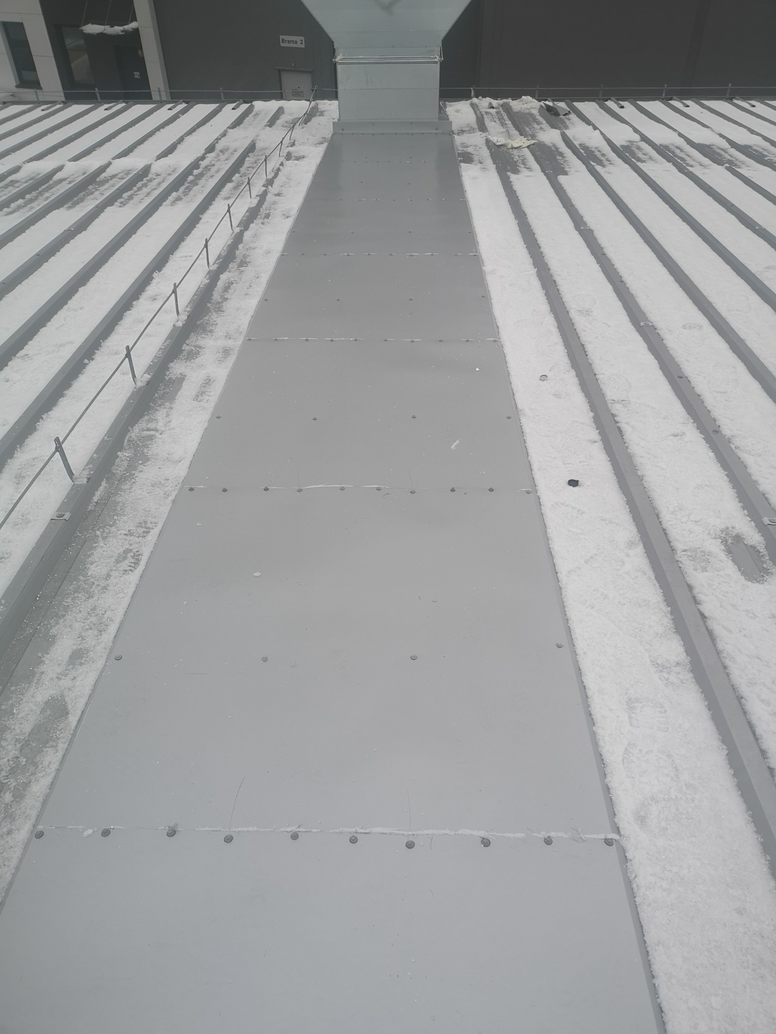 Szary dach z płyt warstwowych pokryty częściowo śniegiem, widoczne łączenia płyt za pomocą nitów.