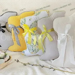 Różnokolorowe poduszki w kształcie królików z kokardami, dekoracyjne pisanki w drewnianej misce i gałązka wierzby na teksturowanym tle.