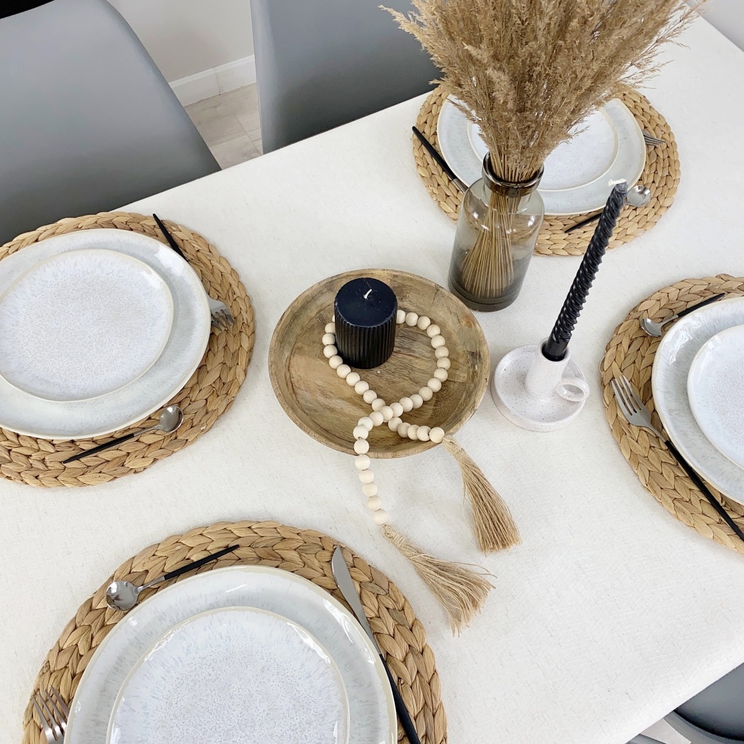 Elegancko nakryty stół z okrągłymi podkładkami z trawy morskiej, białą zastawą, czarnymi sztućcami, świecami i dekoracjami w stylu boho.