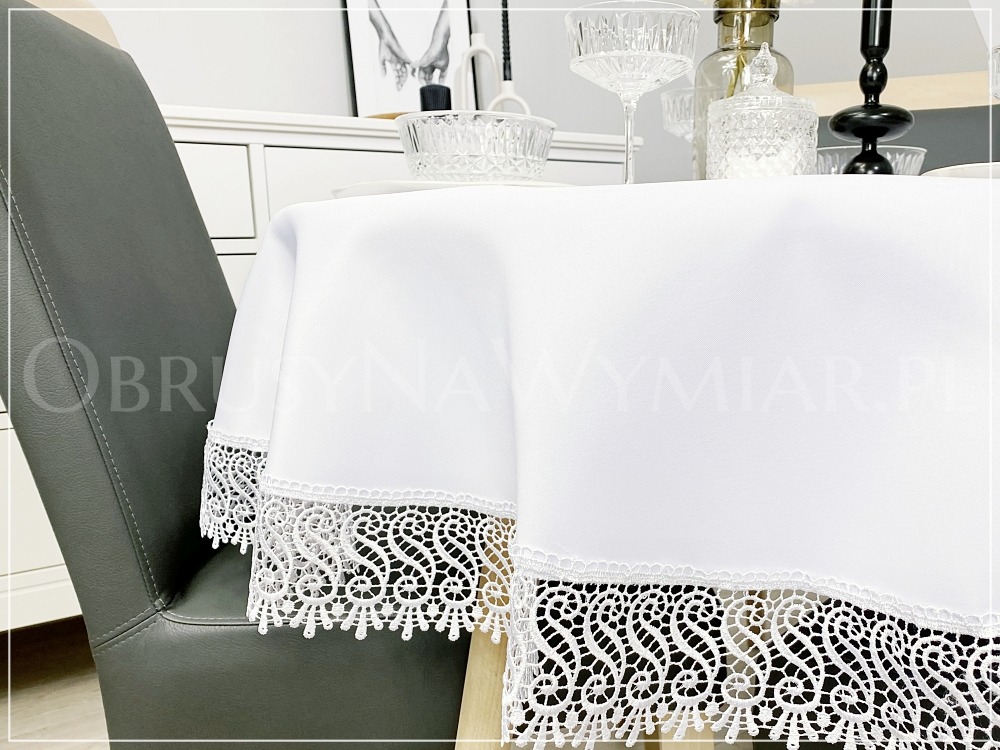 Elegancki biały obrus z koronkowym wykończeniem na stole, z szarą krzesłem w tle i dekoracyjnymi elementami zastawy stołowej.