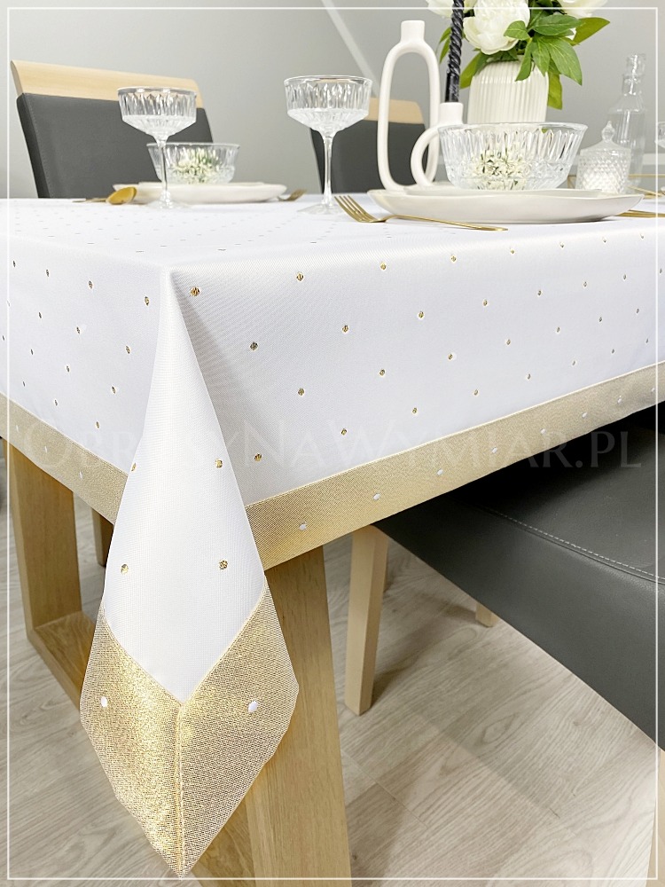 Biały obrus w złote kropki, wykończony złotą obwódką, nakryty na stół z zastawą. Widoczny fragment krzesła i podłogi. Elegancki wystrój stołu.