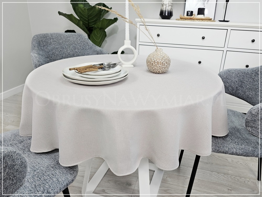 Beżowy, okrągły obrus na stole z zastawą, w tle komoda i roślina. Stonowana kolorystyka, proste krzesła, minimalistyczny wystrój wnętrza.