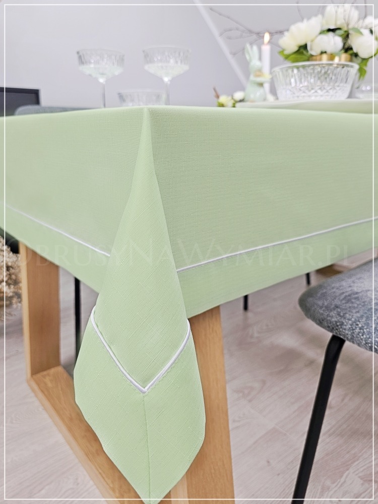 Jasnozielony obrus z białą lamówką na stole z dekoracjami wielkanocnymi w tle. Obrus uszyty na wymiar z dbałością o detale wykończenia.