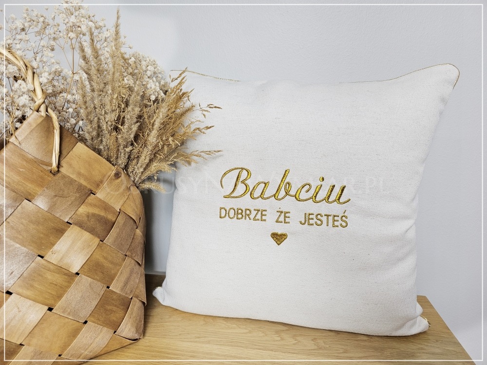 Dekoracyjna, kremowa poduszka z haftowanym napisem 'Babciu, dobrze że jesteś' w złotym kolorze, obok plecionego koszyka z suchymi kwiatami.