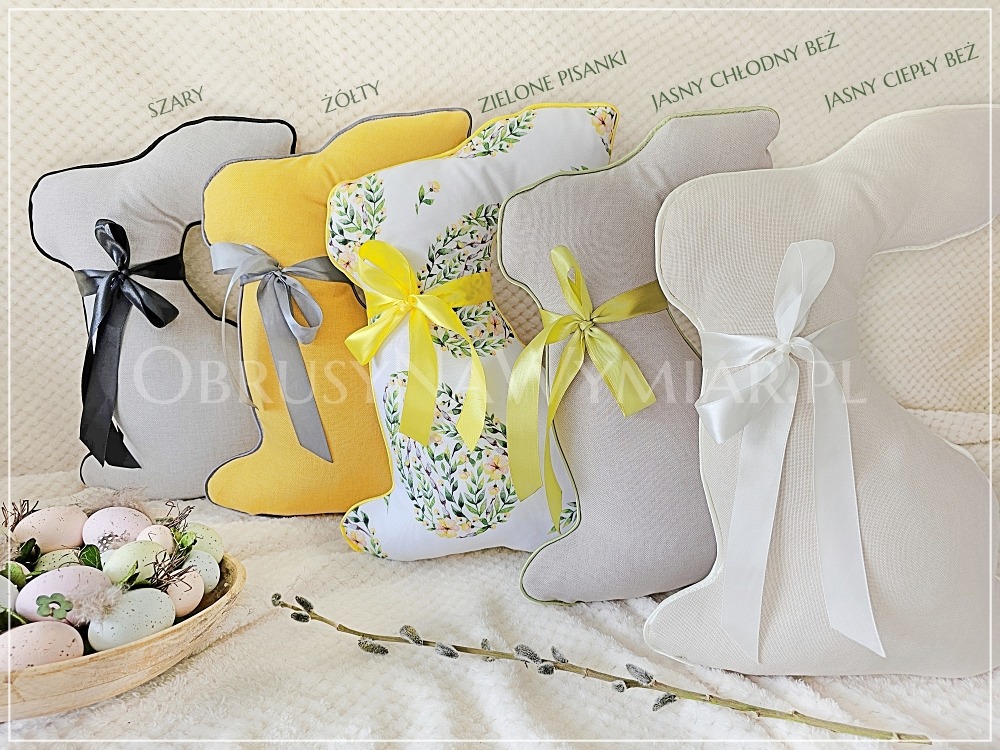 Różnokolorowe poduszki w kształcie królików z kokardami, dekoracyjne pisanki w drewnianej misce i gałązka wierzby na teksturowanym tle.