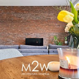 M2W Architektura i Wnętrza - Salon z ceglaną ścianą, szarą sofą modułową, drewnianym stołem i bukietem tulipanów w wazonie. Zapalona świeca na pierwszym planie.