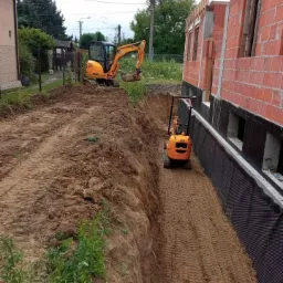 Dwa minikoparki JCB na placu budowy, wykop pod fundamenty domu z cegły, zabezpieczony czarną folią izolacyjną.