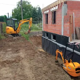 Dwa żółte mini-koparki JCB na placu budowy obok fundamentów domu z czerwonej cegły, zabezpieczonych folią hydroizolacyjną.