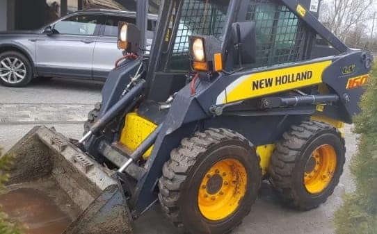 Żółto-czarna miniladowarka New Holland LS160 z łyżką, widoczna na zewnątrz, z zaparkowanym szarym samochodem osobowym w tle.