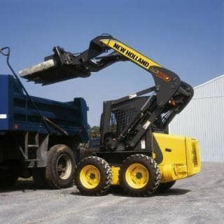 Żółta ładowarka New Holland z podniesioną łyżką, obok niebieskiej wywrotki, na tle szarego budynku.