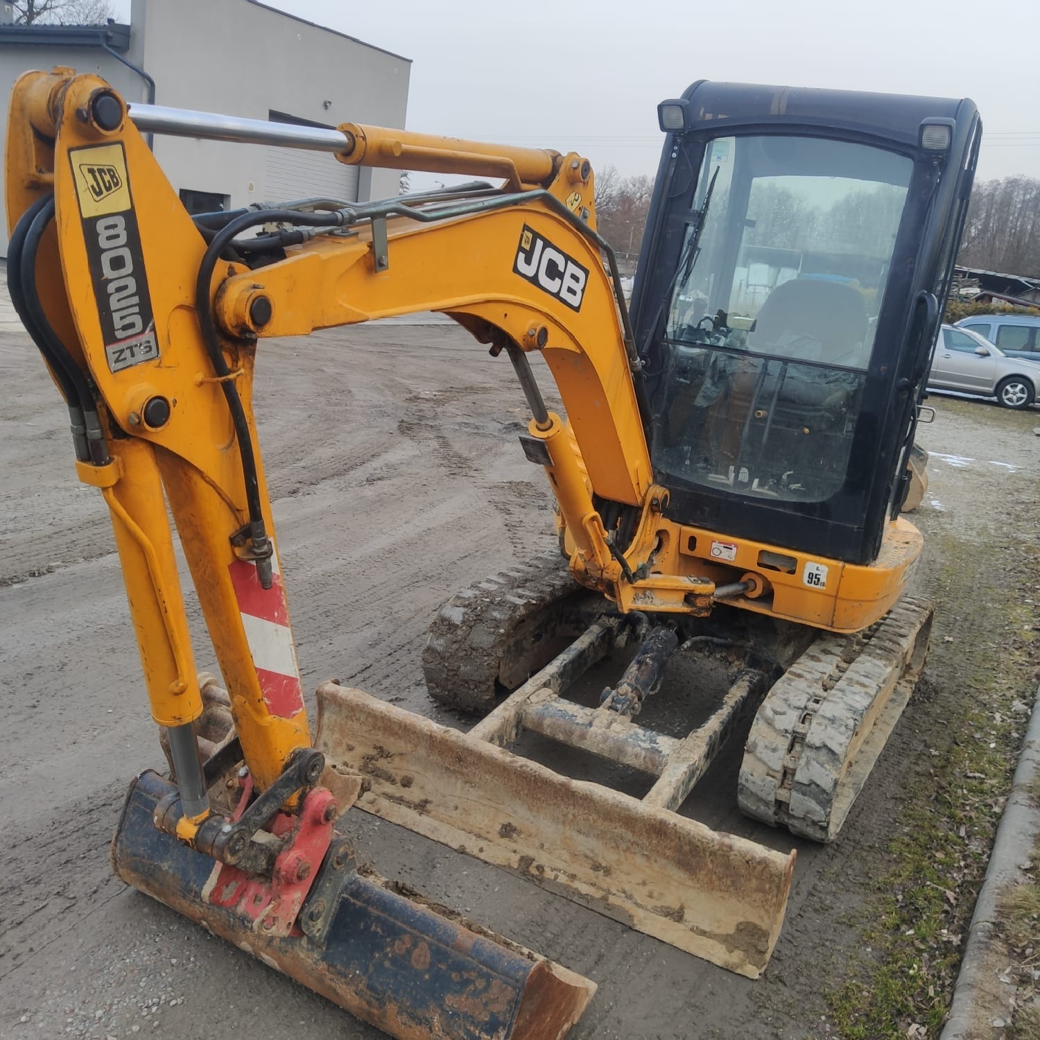 Żółta mini koparka JCB 8025 ZTS z łyżką i lemieszem, stojąca na błotnistym placu budowy, widoczne ślady użytkowania i zabrudzenia.
