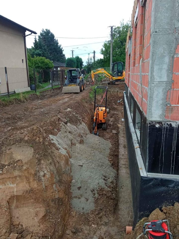 Wykop fundamentów pod budynek z widoczną izolacją pionową, w tle maszyny budowlane: koparki i zagęszczarka stopowa.