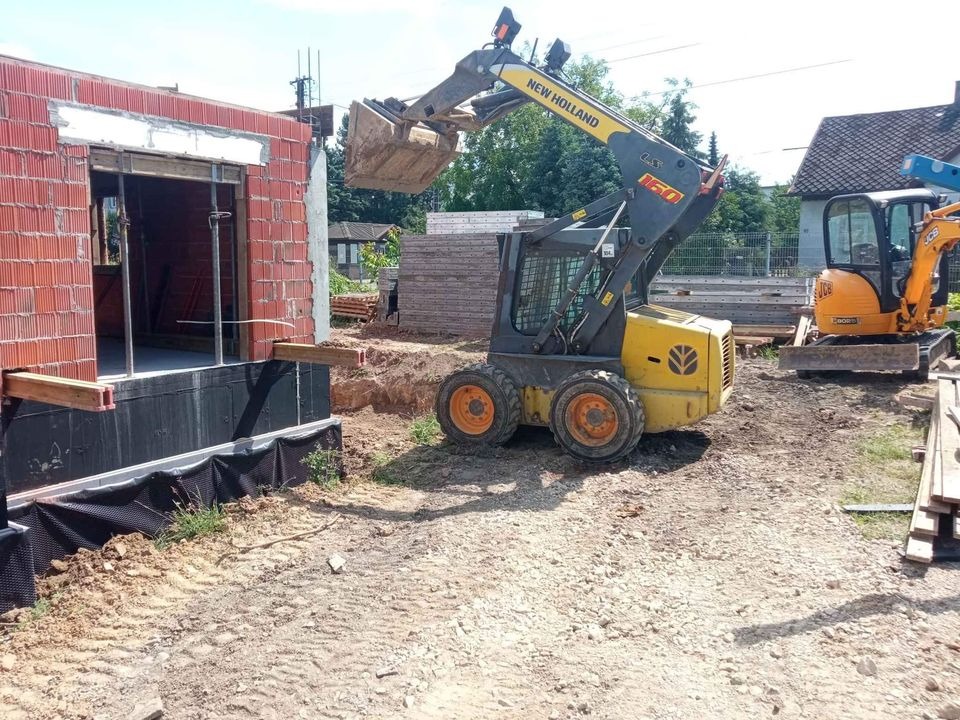 Żółta ładowarka New Holland podnosi ziemię przed budynkiem z czerwonej cegły w trakcie budowy, w tle widoczna koparka JCB.