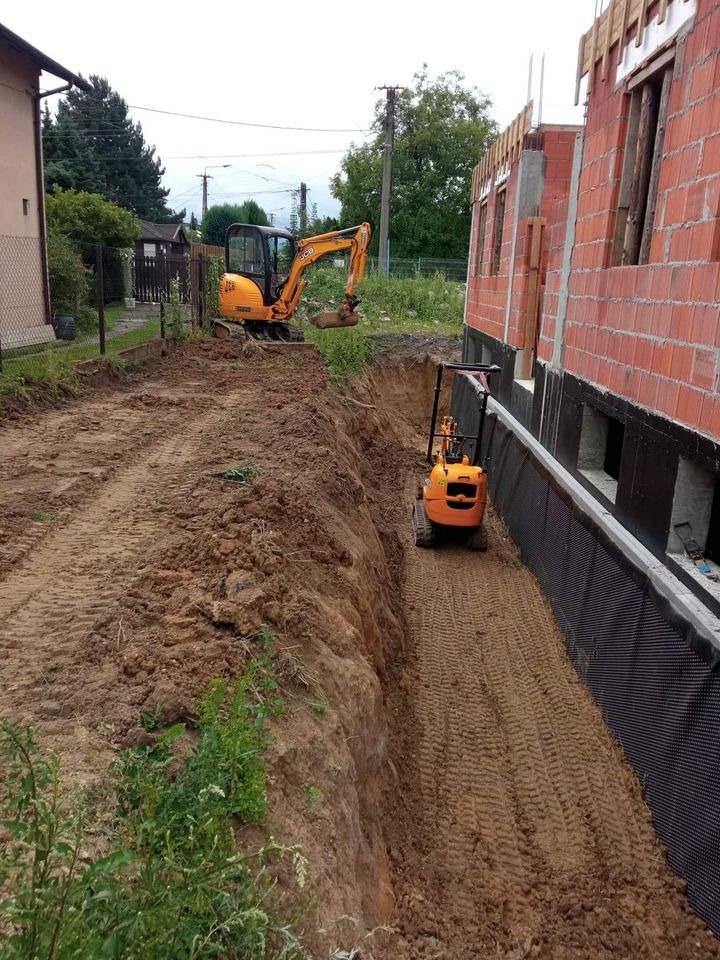Dwa minikoparki JCB na placu budowy, wykop pod fundamenty domu z cegły, zabezpieczony czarną folią izolacyjną.