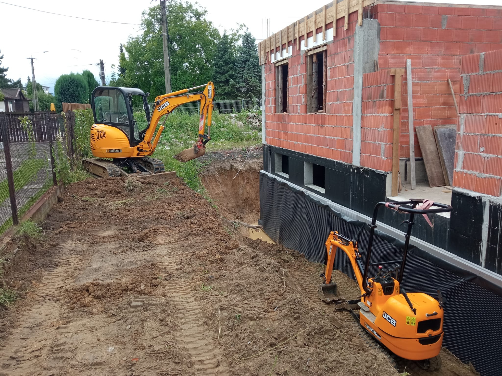 Dwa żółte mini-koparki JCB na placu budowy obok fundamentów domu z czerwonej cegły, zabezpieczonych folią hydroizolacyjną.