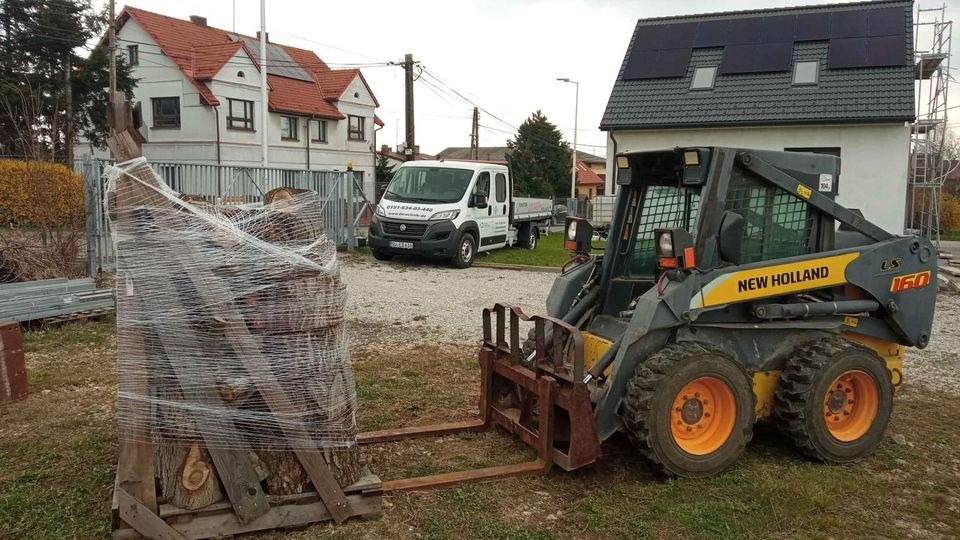 New Holland LS160 ładowarka teleskopowa z paletą drewna opałowego owiniętą folią stretch na placu budowy, w tle budynki mieszkalne i biała furgonetka.
