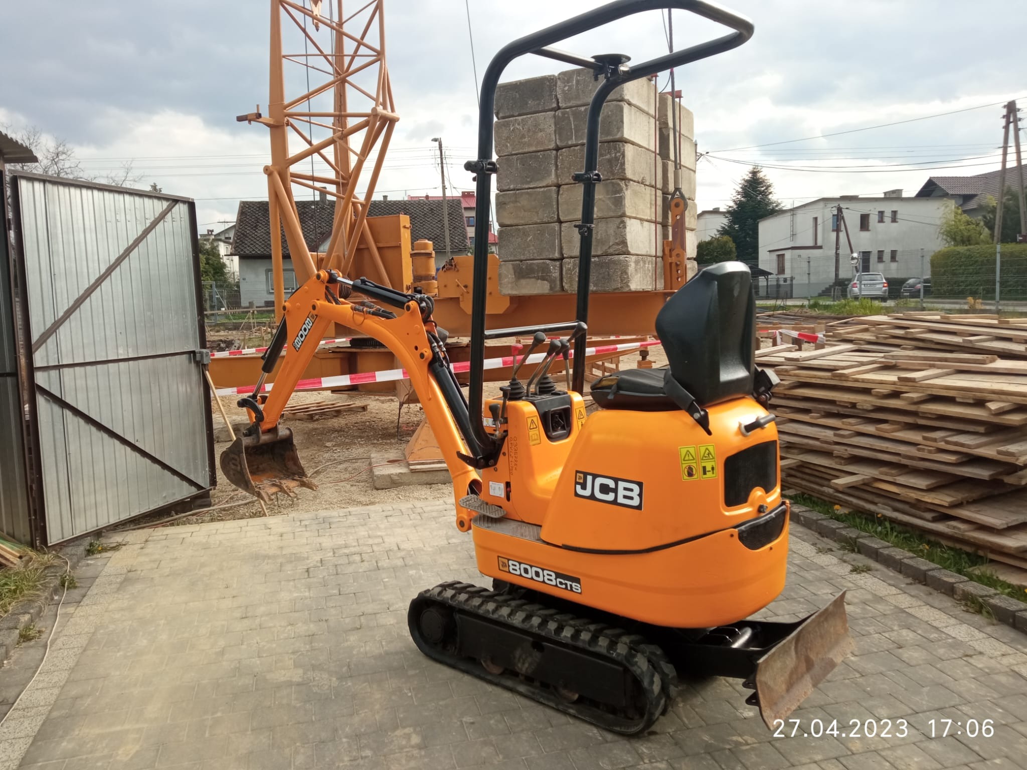 Pomarańczowa mini koparka JCB 8008 CTS na placu budowy w Andrychowie, widoczne deski, żuraw i bloczki betonowe w tle.