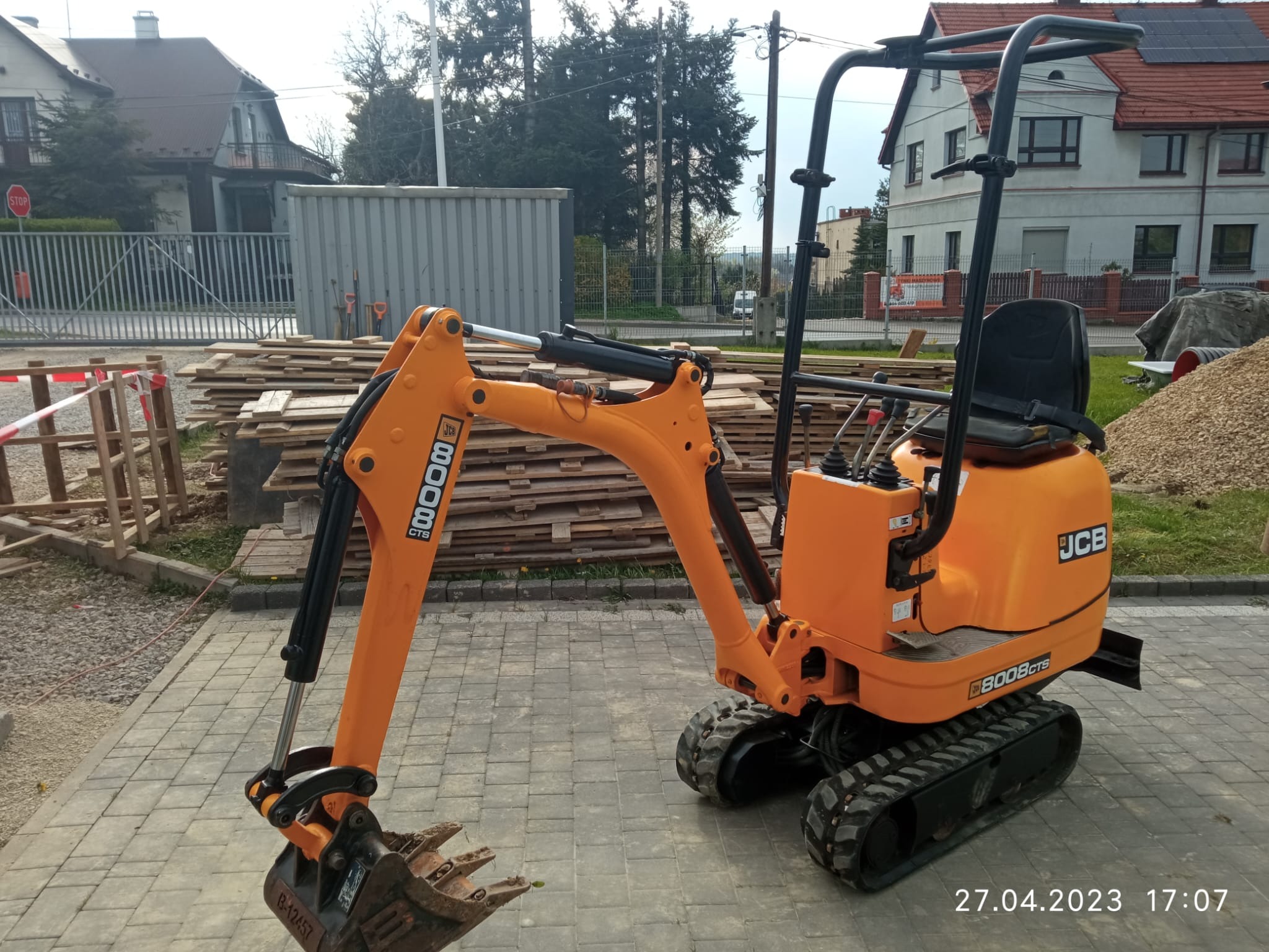 Pomarańczowa mini koparka JCB 8008 CTS zaparkowana na kostce brukowej, w tle sterta drewnianych desek, kontener i budynki mieszkalne.