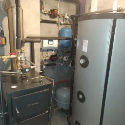 Zasypowy kocioł marki Kamen Eco Design z podłączonym buforem ciepła i zasobnikiem ciepłej wody użytkowej, widoczne liczne rury i zawory w pomieszczeniu kotłowni wykończonym szarą płytką.