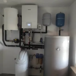 Kompletna instalacja grzewcza w kotłowni: biały wiszący kocioł gazowy Bosch z wyświetlaczem, zbiornik buforowy Galmet, dwa zbiorniki wyrównawcze, zmiękczacz wody USTM oraz liczne rury i zawory.