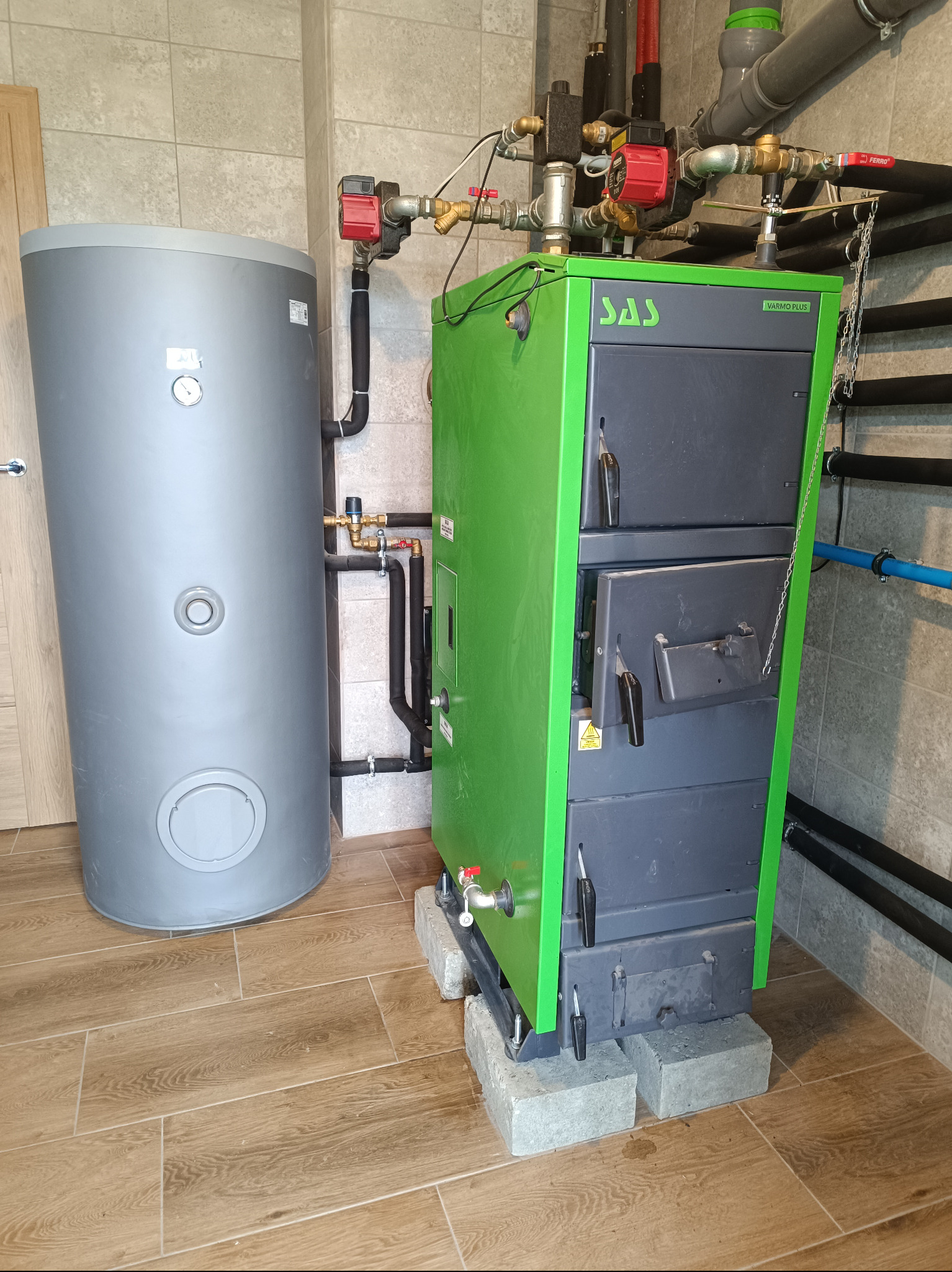 Zielono-szary piec SAS Warmo Plus na betonowych bloczkach, obok szary zasobnik ciepłej wody, instalacja grzewcza z pompą obiegową i rurami w tle.