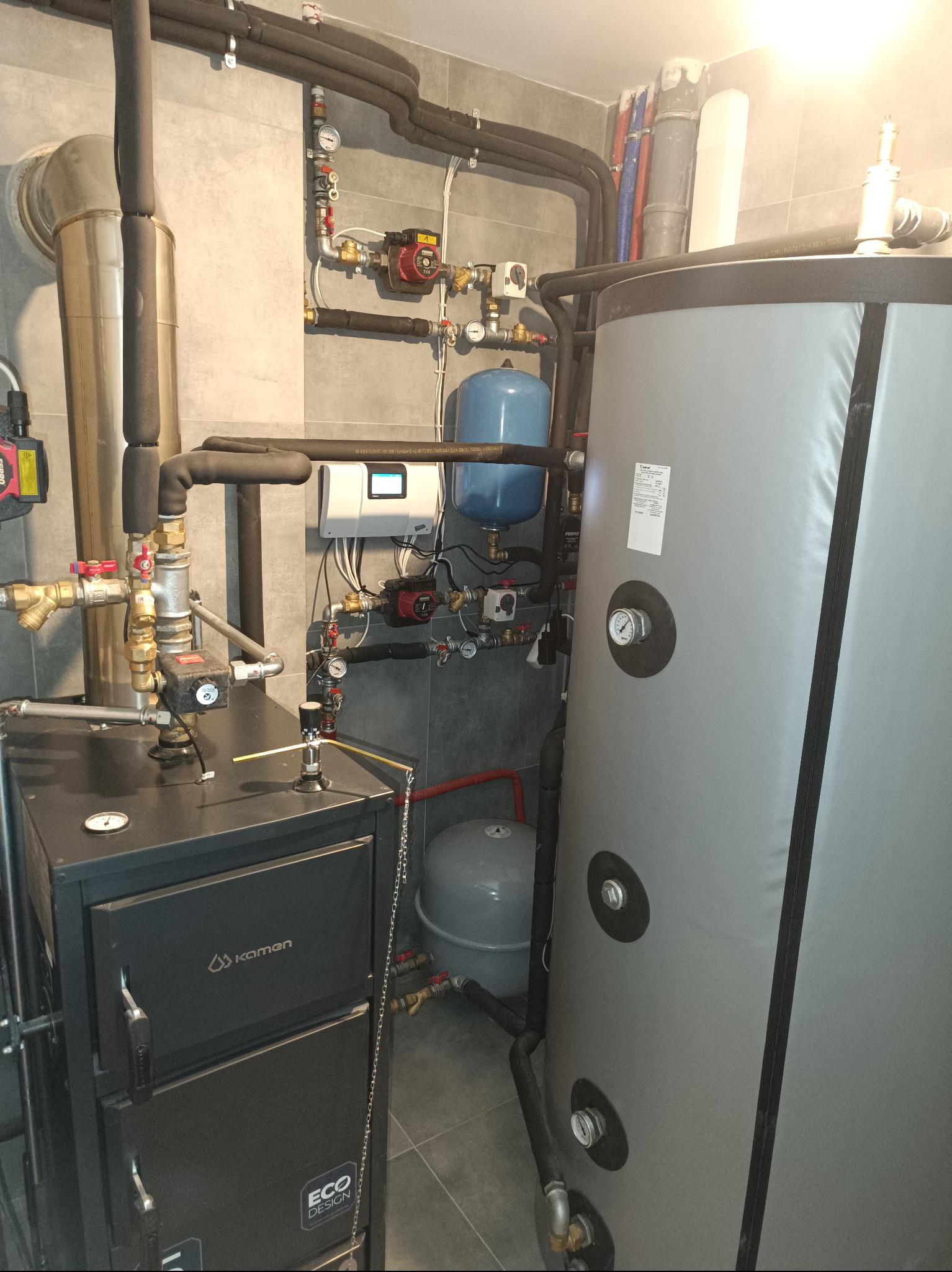 Zasypowy kocioł marki Kamen Eco Design z podłączonym buforem ciepła i zasobnikiem ciepłej wody użytkowej, widoczne liczne rury i zawory w pomieszczeniu kotłowni wykończonym szarą płytką.