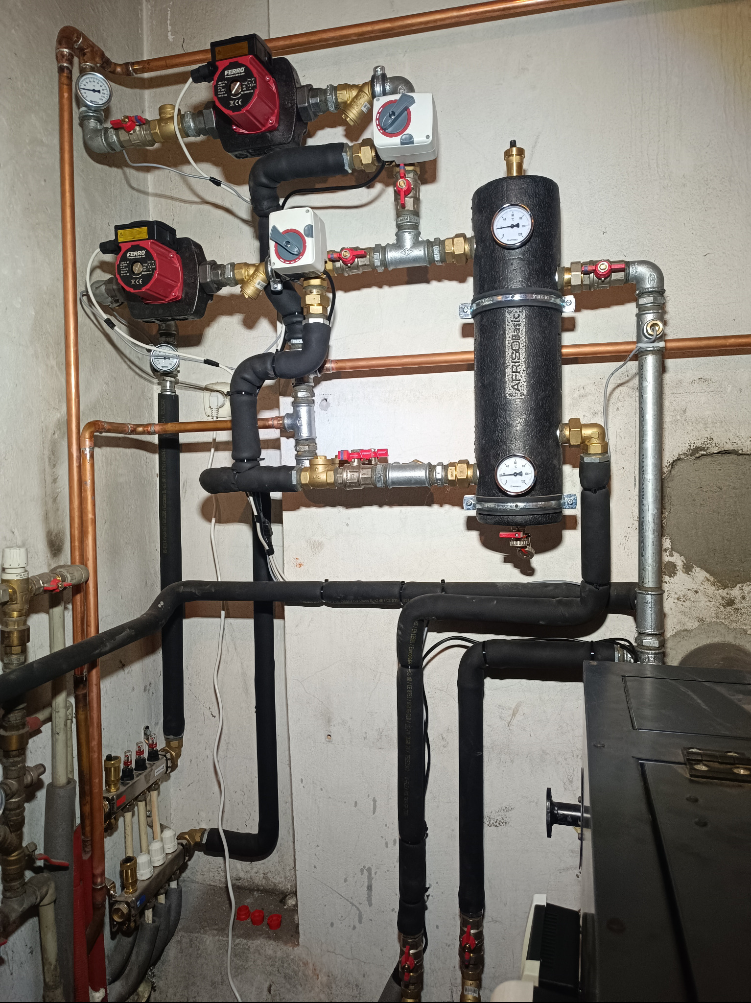 Złożona instalacja grzewcza z pompami obiegowymi Ferro, zbiornikiem AFRISO Basic, rurami miedzianymi i czarnymi, izolowanymi przewodami, manometrami i zaworami, zamontowana na tle surowej ściany.