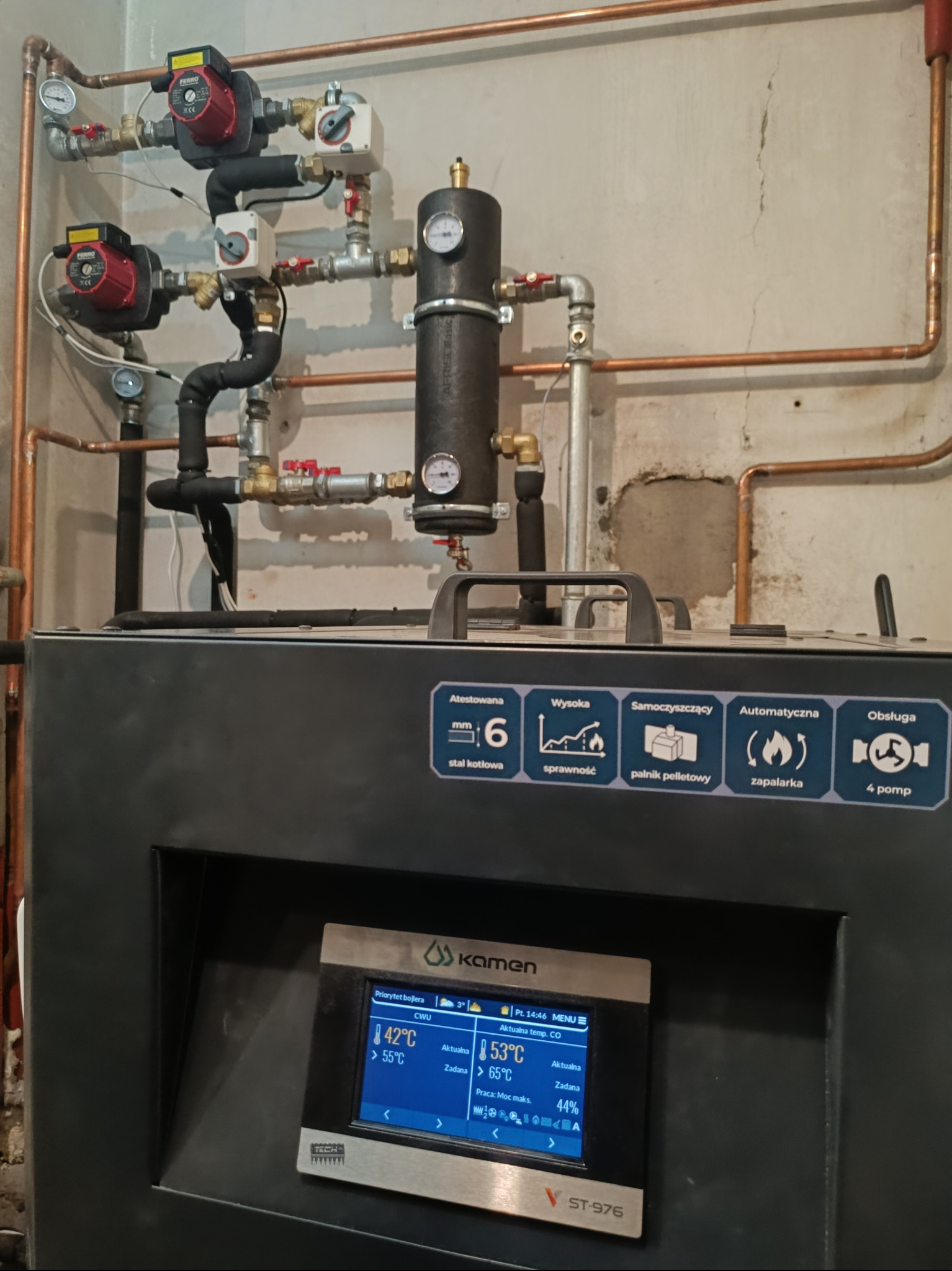 Nowoczesny piec z automatycznym podajnikiem pelletu, widoczne podłączenia hydrauliczne z zaworami i pompami obiegowymi oraz sterownik z wyświetlanymi temperaturami pracy.