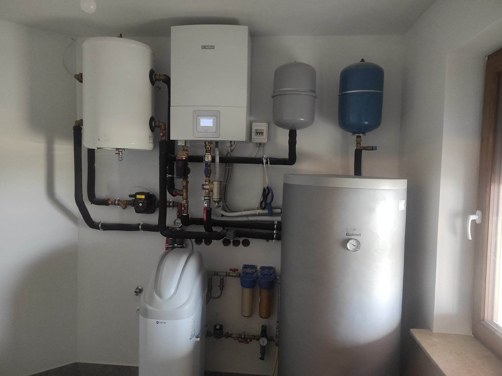 Kompletna instalacja grzewcza w kotłowni: biały wiszący kocioł gazowy Bosch z wyświetlaczem, zbiornik buforowy Galmet, dwa zbiorniki wyrównawcze, zmiękczacz wody USTM oraz liczne rury i zawory.