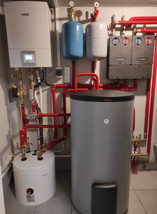 Zestaw urządzeń grzewczych marki Bosch z kotłem, zasobnikiem ciepłej wody, naczyniami wzbiorczymi i rozdzielaczem, połączonych rurami w kolorze czerwonym.