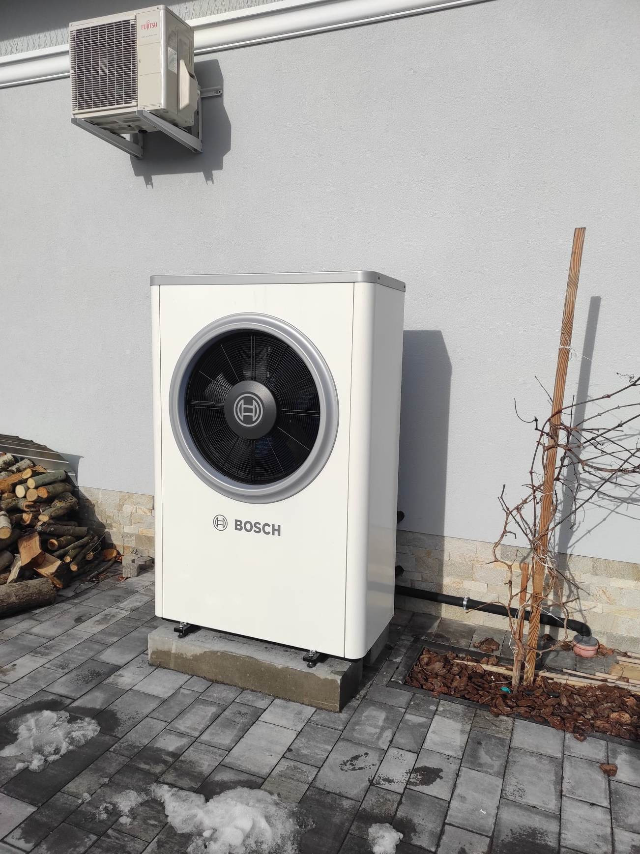 Biała pompa ciepła Bosch na betonowym fundamencie, z widocznym klimatyzatorem Fujitsu na szarej ścianie budynku. W tle stos drewna i fragment uschniętej rośliny.