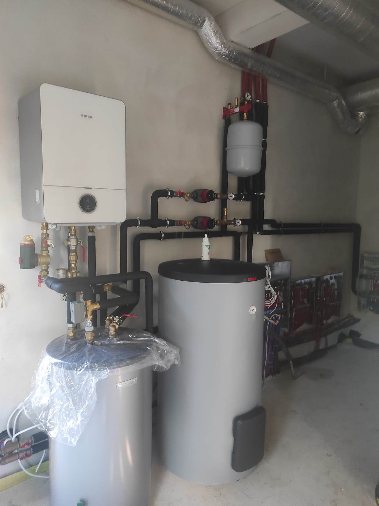 Nowoczesna instalacja grzewcza z piecem gazowym marki Bosch, dwoma zbiornikami akumulacyjnymi, rurami z czarnymi izolacjami i zaworami, widoczne przewody wentylacyjne pod sufitem.