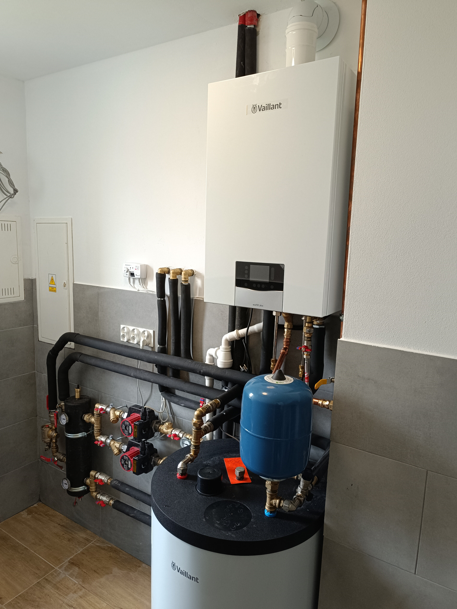 Nowoczesny, biały kocioł gazowy Vaillant wiszący na ścianie, z widocznymi rurami, zaworami i zbiornikiem wyrównawczym w kolorze niebieskim, zainstalowany w pomieszczeniu z szarą glazurą i brązowymi...