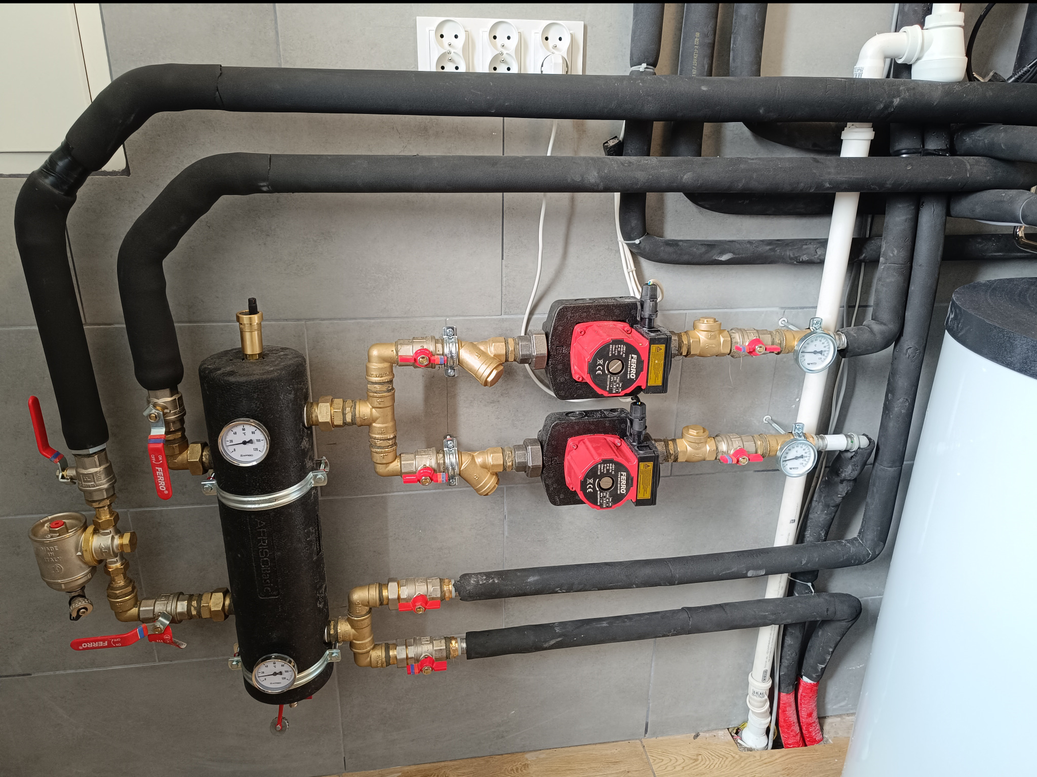 Złożona instalacja grzewcza z czarnymi rurami izolowanymi, dwoma pompami obiegowymi FERRO z czerwonymi obudowami, zbiornikiem wyrównawczym AFRISOBasic z manometrami i licznymi zaworami odcinającymi...