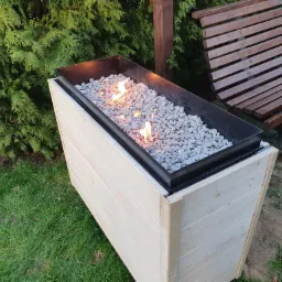 Ognisko ogrodowe w drewnianej obudowie, wypełnione kamieniami, z widocznymi płomieniami, na tle zieleni i ławki parkowej.