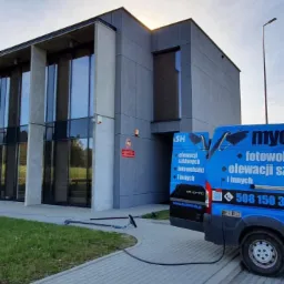 Nowoczesny budynek biurowy z dużymi oknami, obok którego stoi niebieski samochód dostawczy z reklamą firmy świadczącej usługi mycia elewacji i okien, oraz rozłożone narzędzia do mycia.