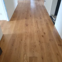 Świeżo położona, lakierowana podłoga z paneli drewnianych w jasnym odcieniu dębu, widok w perspektywie korytarza, z widocznymi drobnymi niedoskonałościami montażu przy ścianach.