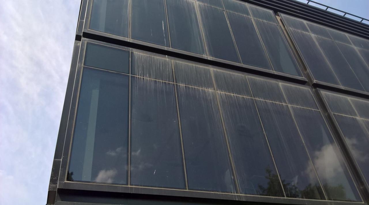 Zewnętrzna ściana biurowca z bardzo brudnymi oknami, widoczne zacieki i osady, niebo w tle.