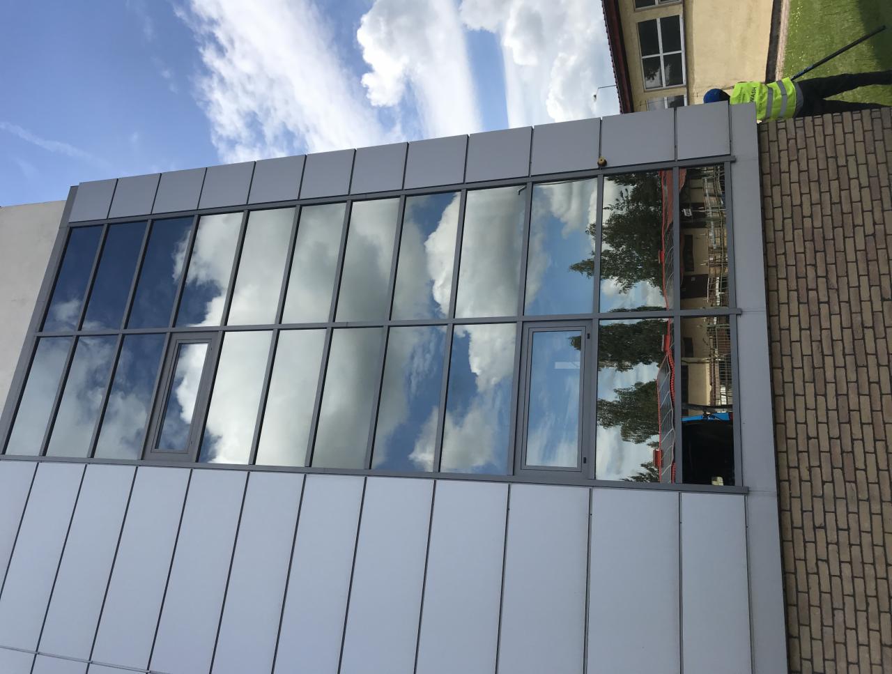 Pracownik w odblaskowej kamizelce myje okna na wysokości w budynku biurowym, w szybach odbija się błękitne niebo z chmurami.