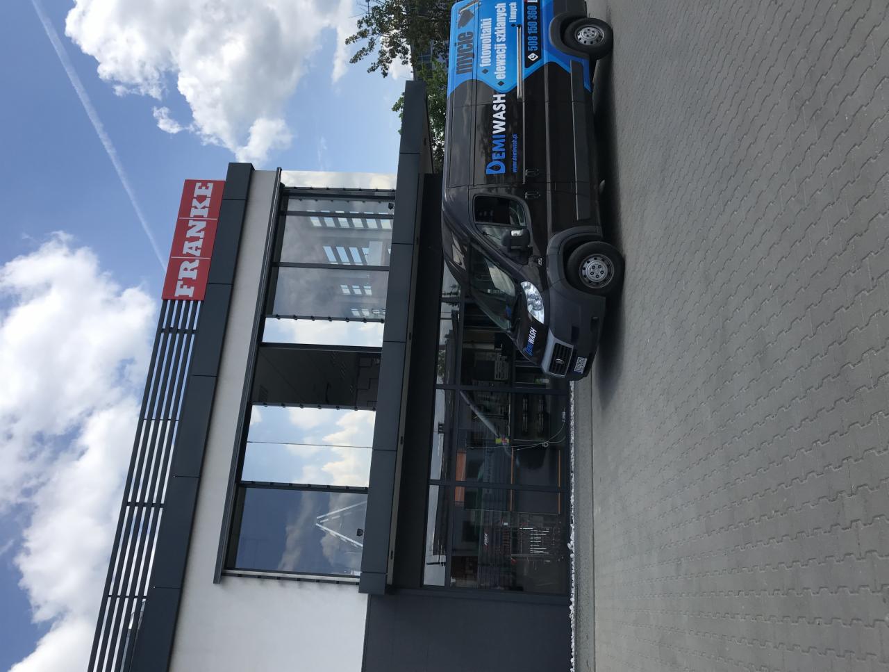 Czarny samochód dostawczy firmy Demiwash z reklamą mycia fotowoltaiki i elewacji szklanych zaparkowany przed budynkiem z dużymi oknami i logo Franke.