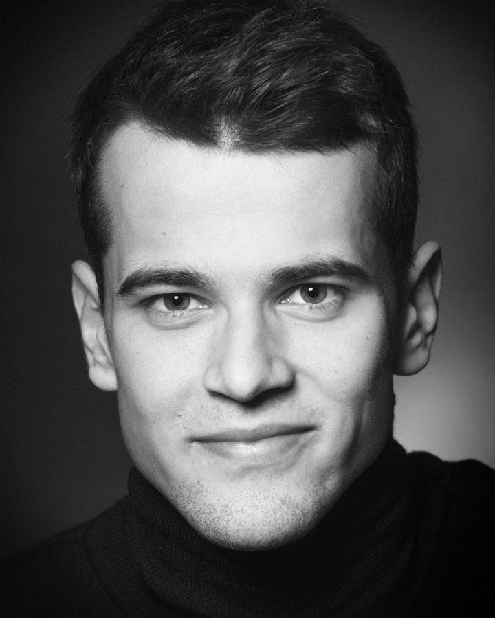 Czarno-biały portret młodego mężczyzny z krótkimi włosami, ubranego w czarny golf, z delikatnym uśmiechem i łagodnym spojrzeniem, na ciemnym tle.